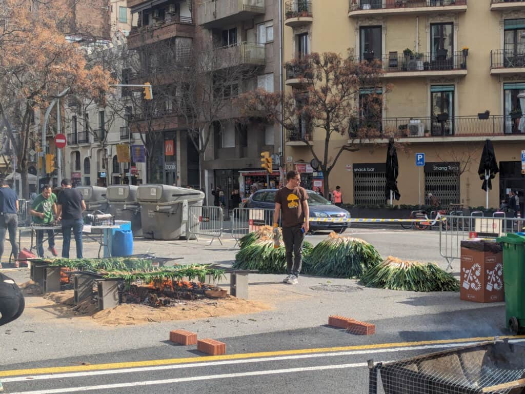 Spanish Food Culture: Discover Spain Through Its Traditional Cuisine An outdoor street scene in Barcelona during a traditional calçotada celebration. Large bundles of calçots, a type of Catalan green onion, are stacked beside open grills where they are being roasted over fire. People work around the cooking area while apartment buildings and city streets form the backdrop, highlighting a beloved culinary tradition of Catalonia and Spanish food culture.