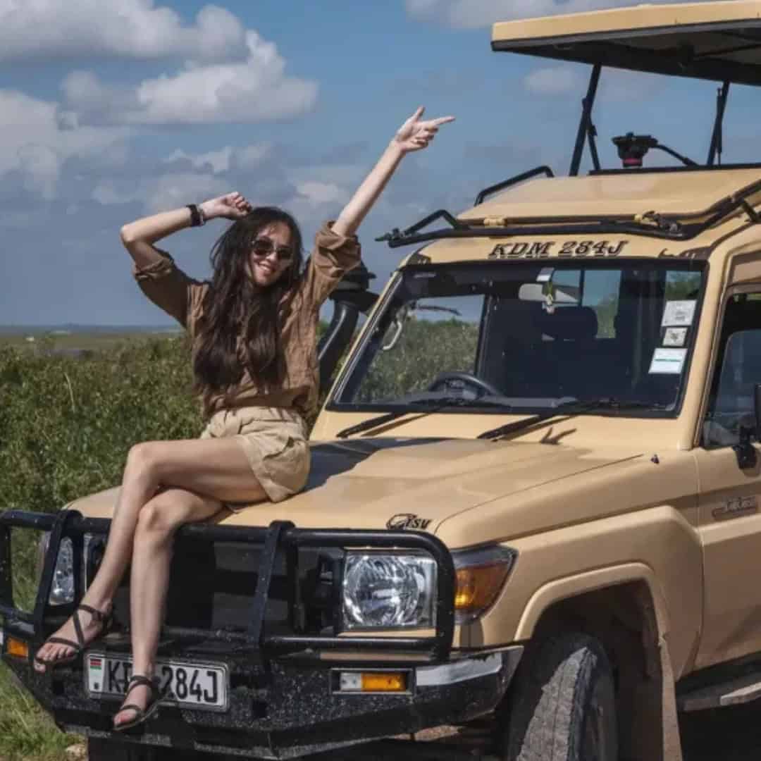 How to Plan a Visit to Ngorongoro Crater? My Personal Safari Experience A woman wearing sunglasses, a brown shirt, and beige shorts sits on the hood of a beige safari vehicle with one arm raised and pointing toward the sky. The off-road vehicle is parked in an open grassy savanna under a blue sky with scattered clouds, creating a joyful and adventurous safari scene.