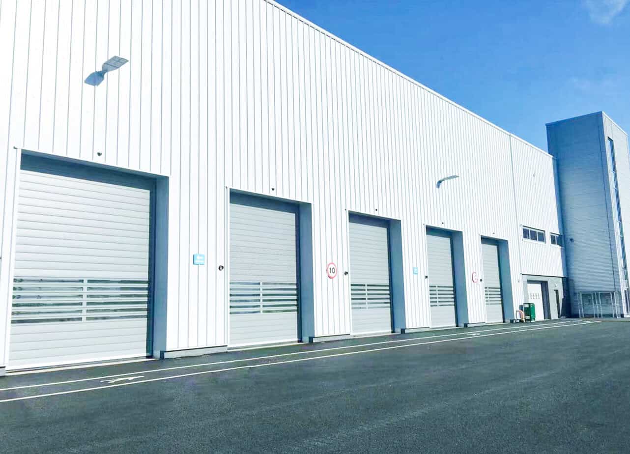 Steel industrial warehouse with multiple roll-up garage doors and bright exterior lighting under a clear blue sky.