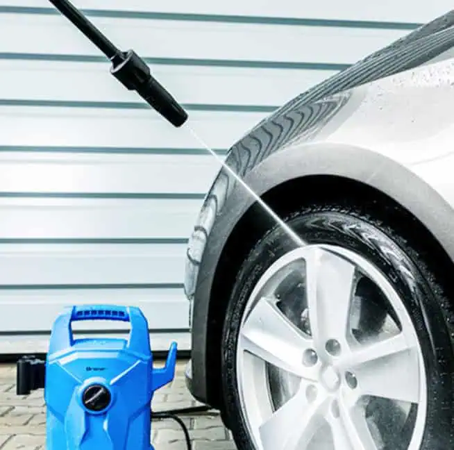 High-pressure car wash using a professional pressure washer cleaning a silver vehicle at an outdoor station.