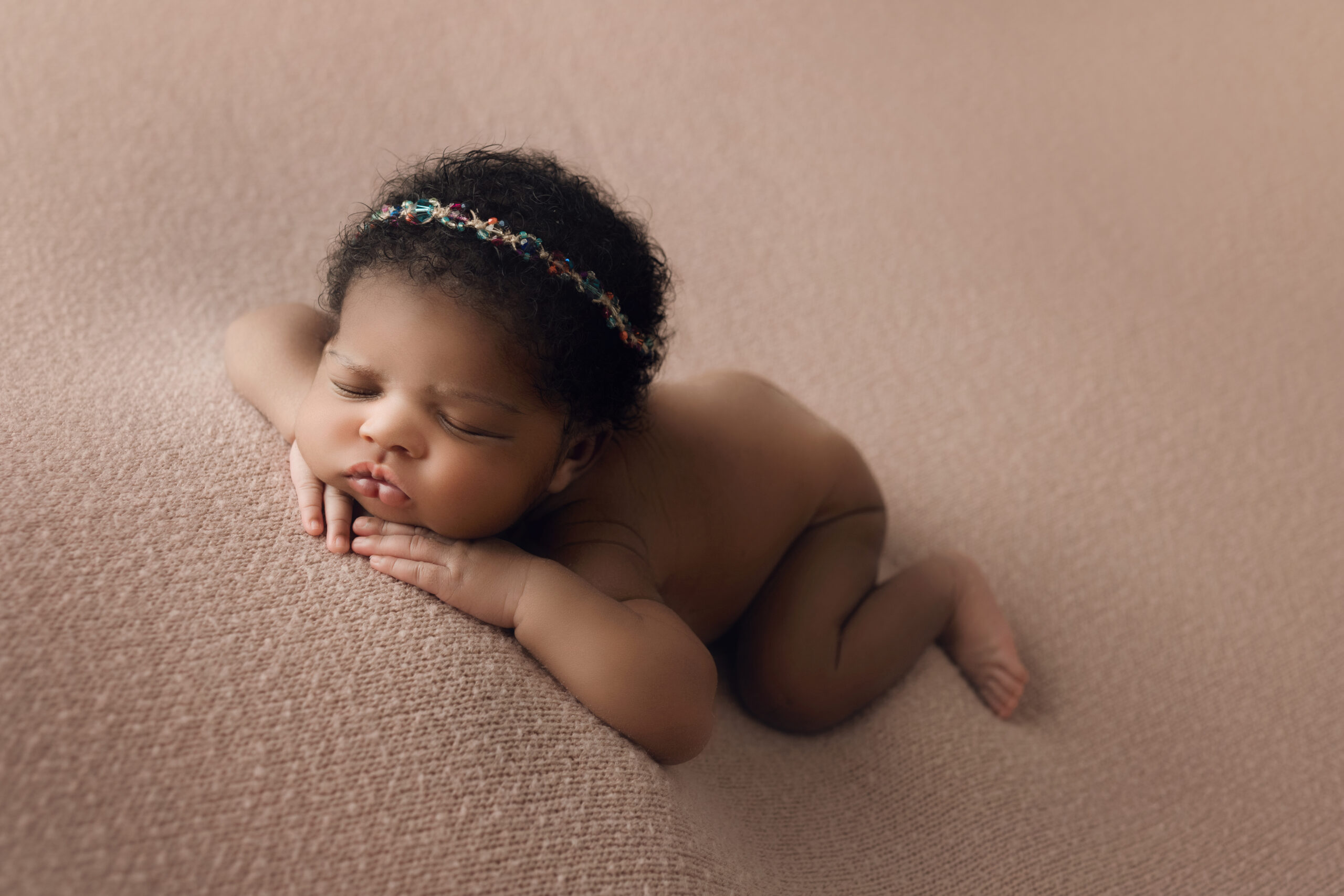 sleepy baby girl face forward chin on hands wearing a princess headband on a pink blanket