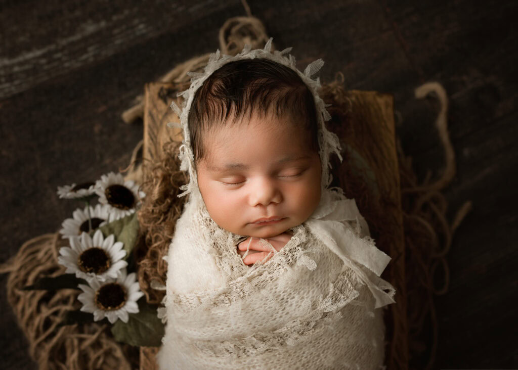 Newborn baby girl sleeping in a wooden prop wrapped in cream and matching bonnet with flowers
