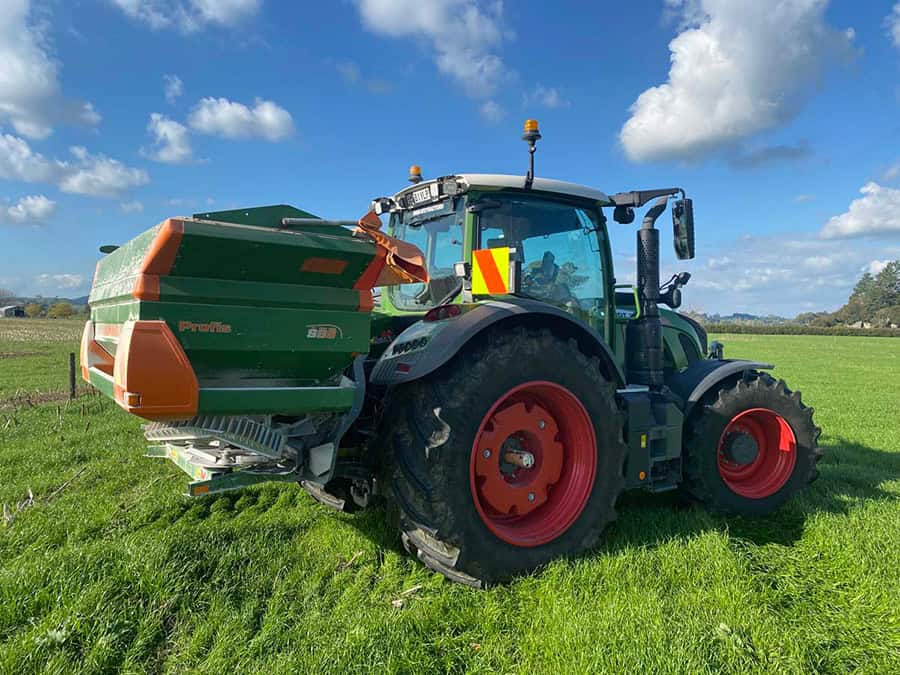 Efficient farming tractor with fertiliser spreader on green field under blue sky.
