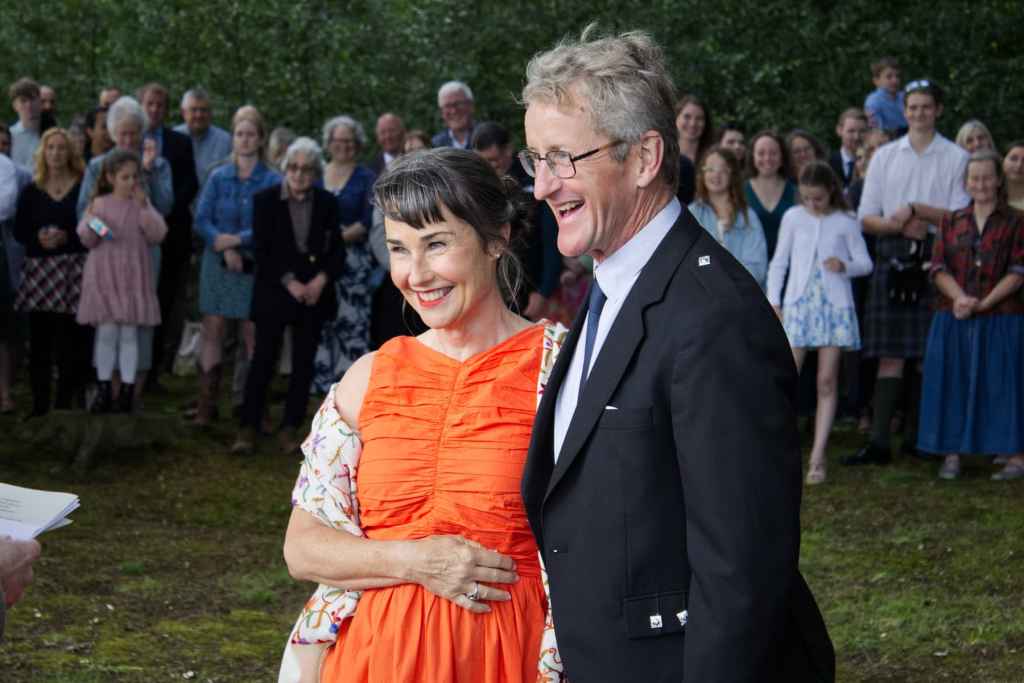 Smiling couple at outdoor event with crowd in background, summer setting.