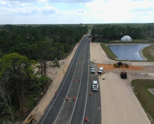 Flexible asphalt road construction with traffic cones and construction vehicles in progress near a water body, surrounded by lush forest and under construction at a site development project.