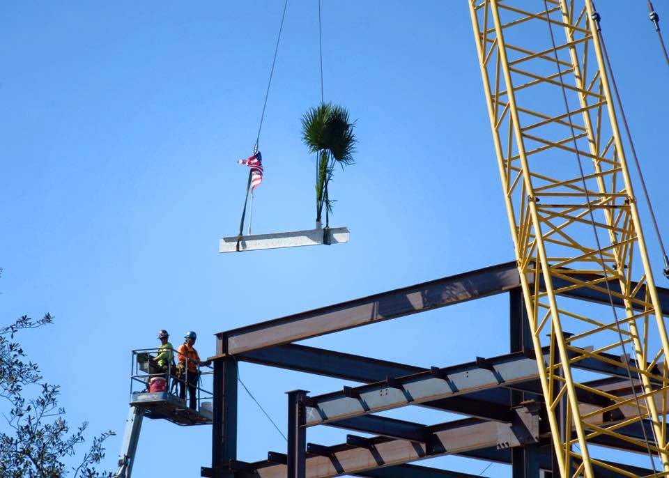 Aerial view of construction workers on a lift installing a palm tree on a building framework with a yellow crane, part of Florida site development projects.