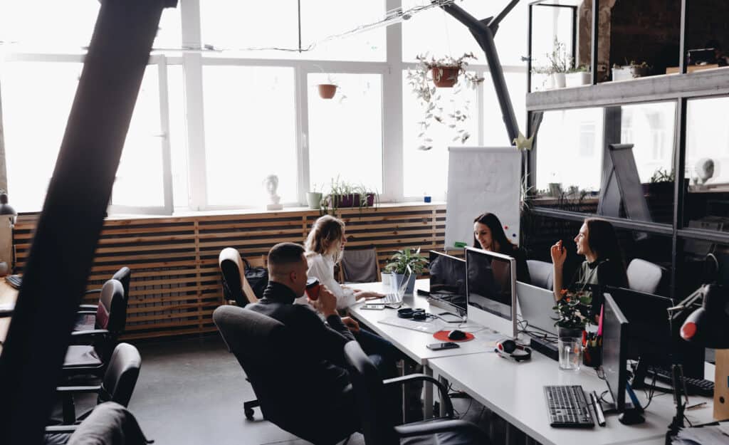 Modern office workspace with team members collaborating, multiple computer monitors, natural light, plants, and creative environment for innovative work and team synergy.