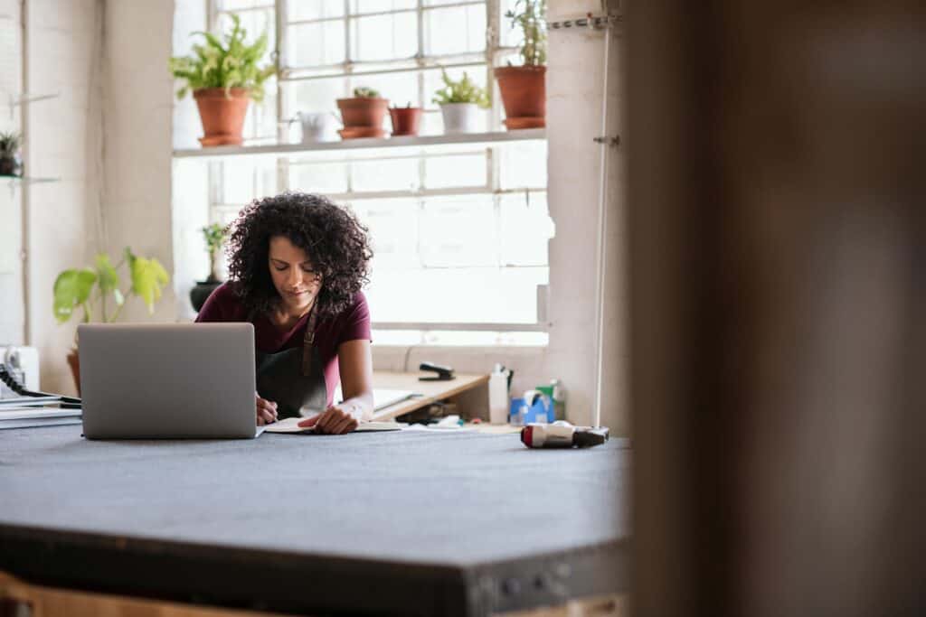 Efficient woman working on her laptop in a bright, modern workshop or co-working space with plants and large windows for optimal productivity and creativity.