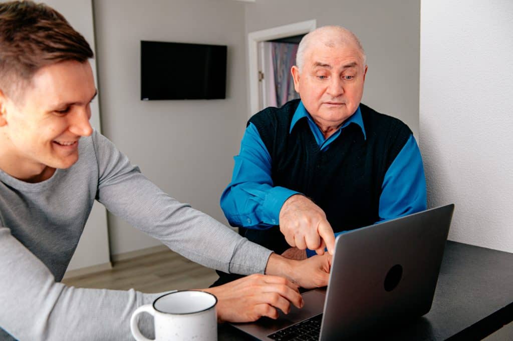 An older man receives help on his laptop computer from a younger man