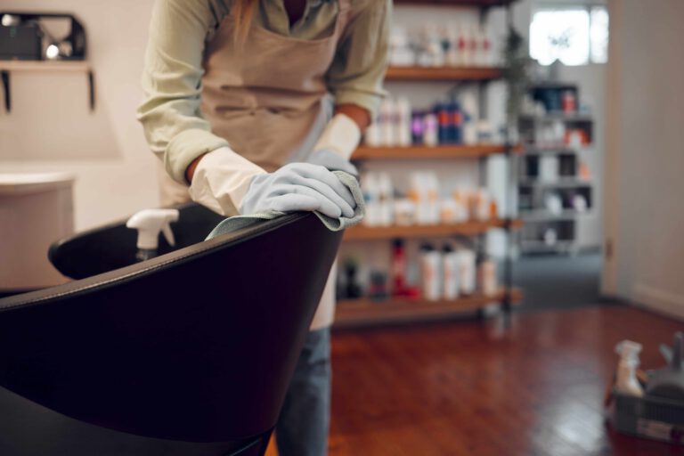 A cleaner wearing gloves and an apron is cleaning a black chair in a well-organized, modern interior with shelves of supplies in the background.