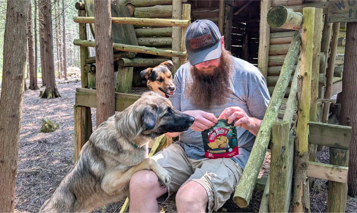 Homem abrindo um pacote de guloseimas com dois cães ao seu lado | Feito no Canadá: nossas guloseimas favoritas de cães e gatos canadenses