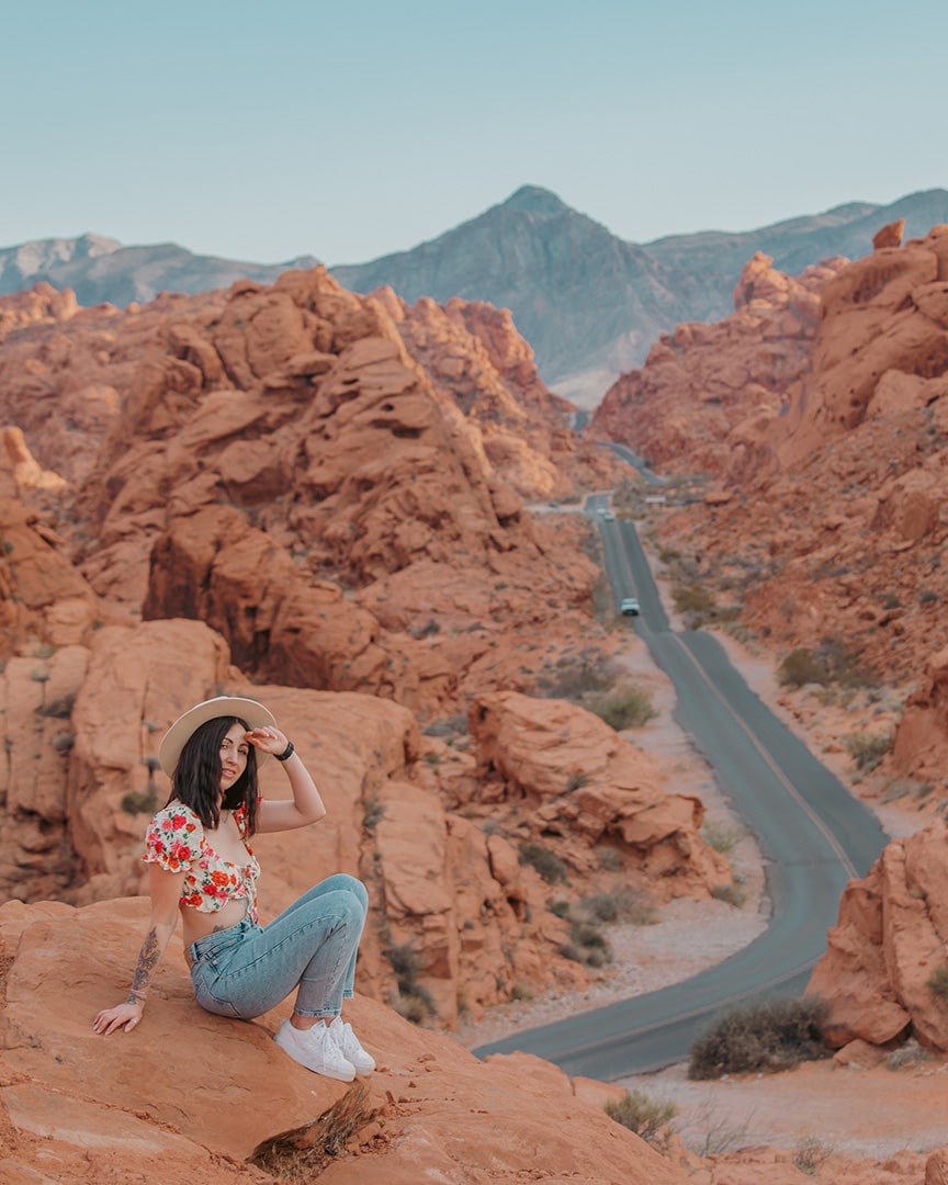 A young woman sits on a large red rock formation in a desert landscape, wearing light blue jeans, a white hat, and a red floral crop top. She’s looking off into the distance with one hand shading her eyes. Behind her, a winding road snakes through dramatic red sandstone canyons, and mountains rise in the background under a clear blue sky.