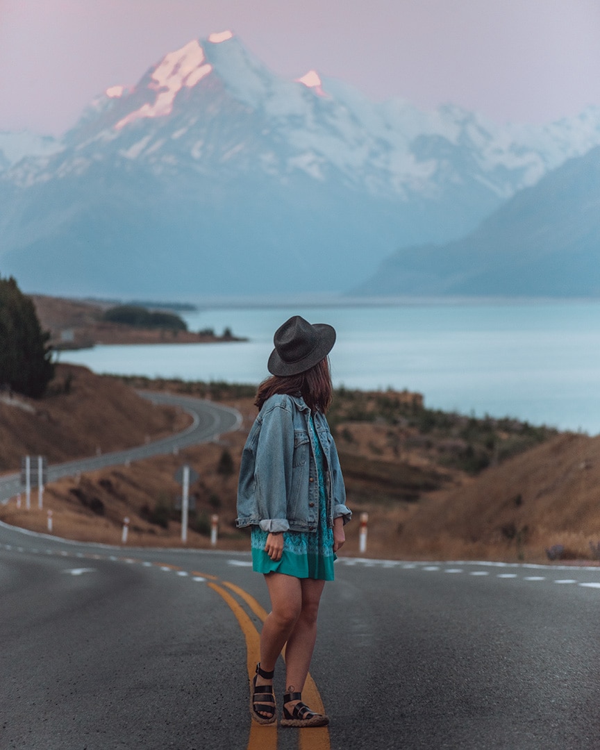 A girl walking down a road next to a turquoise lake. At the top of the lake is a tall snowy mountain. The sky is purple from sunset. Lake Pukaki & Mount Cook, NZ South Island Road Trip.