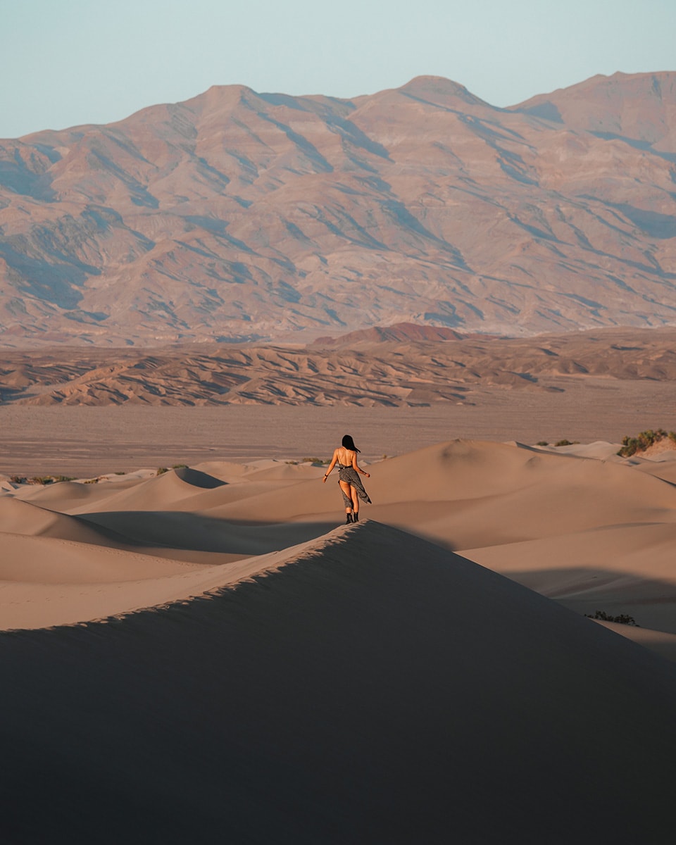 A woman walks barefoot along the top of a golden sand dune at sunset, surrounded by a vast desert and distant mountains. 