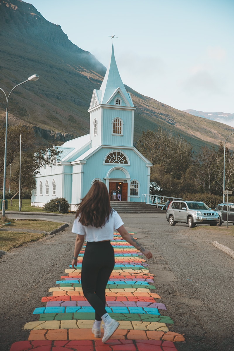 A woman walks along a colorful rainbow-painted brick path toward a pale blue church with a mountain backdrop in a quiet Icelandic town.