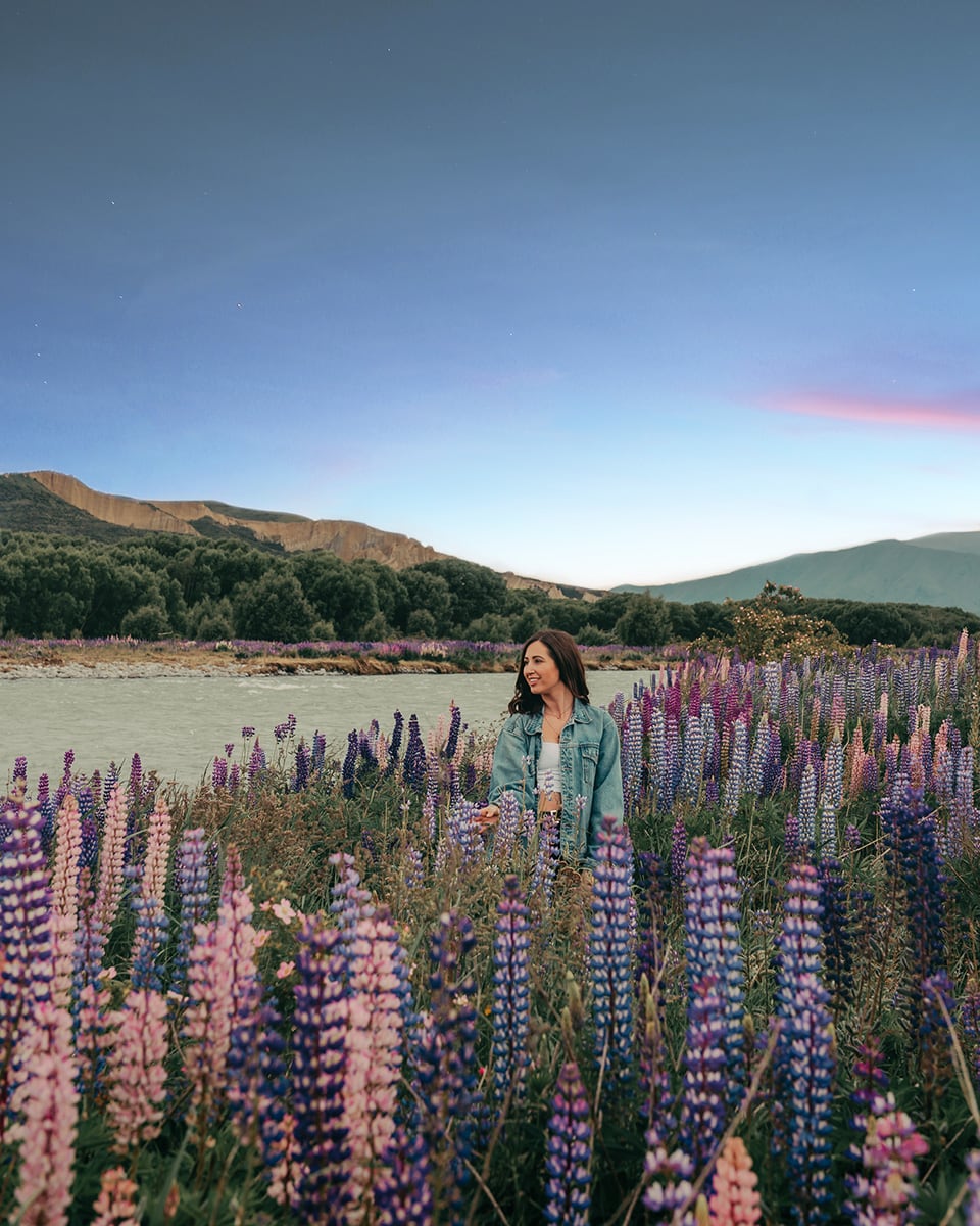 A woman stands in the middle of a vibrant field of lupins in full bloom, surrounded by tall flowers in shades of purple, pink, and lilac. She’s smiling and wearing a denim jacket over a white top, with long dark hair falling over her shoulders. In the background, a gentle river flows past the field, and beyond it are green trees, hills, and a soft evening sky fading into blue and pale pink.