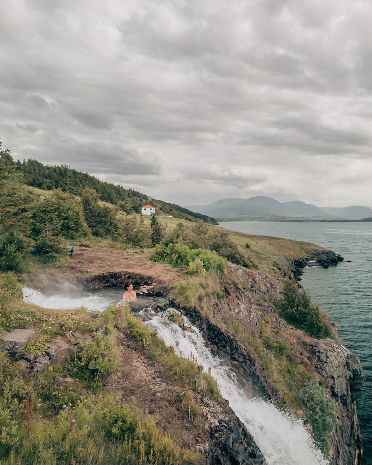 A woman relaxes in a small natural hot spring beside a waterfall, perched on a grassy cliff overlooking the ocean in Iceland. A white house sits in the distance under a cloudy sky.