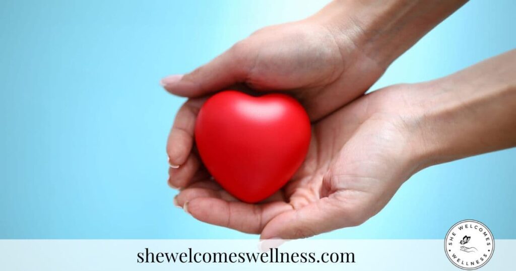Hands gently cradle a red heart against a blue background, conveying care and warmth. The text "shewelcomeswellness.com" is visible below.