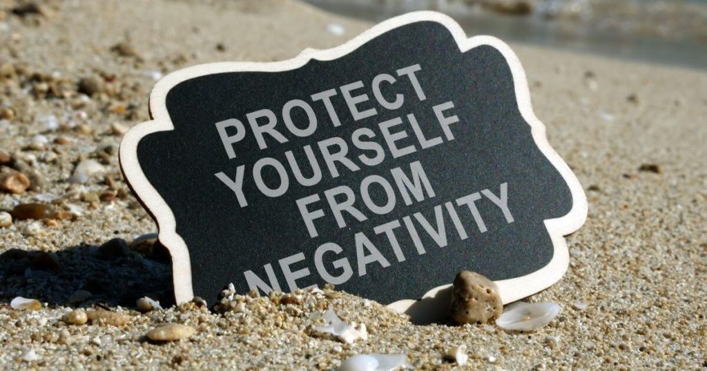 black sign on sand, white words: protect yourself from negativity