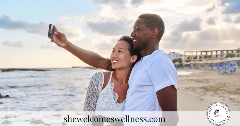 A smiling couple takes a selfie on a beach at sunset, exuding warmth and joy. Waves gently crash behind them under a partly cloudy sky.