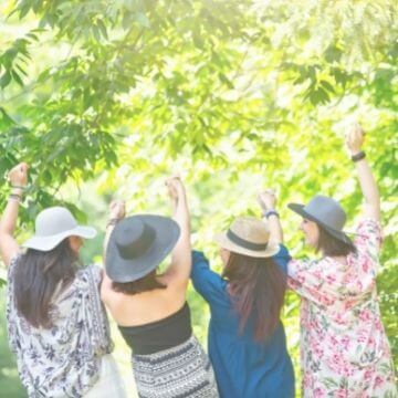 Four women in hats with hands raised, backs to camera, greenery and sun in front