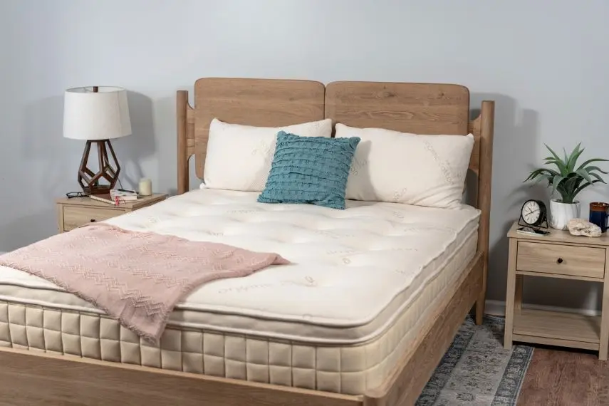 A cozy bedroom featuring a bed with a Naturepedic organic mattress, wooden headboard. The bed has a white mattress, cream pillows, a teal cushion, and a pink throw.
