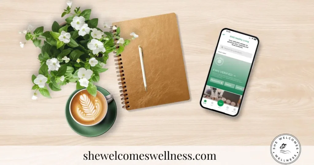 A cozy setup features a cappuccino with latte art, a green cup, a spiral notebook, a pen, a smartphone with green EWG app screen, and white flowers on a wooden table.