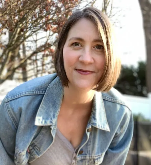 Tina, a woman with short brown hair, smiles warmly at the camera wearing a denim jacket. The background features a tree with brown leaves.