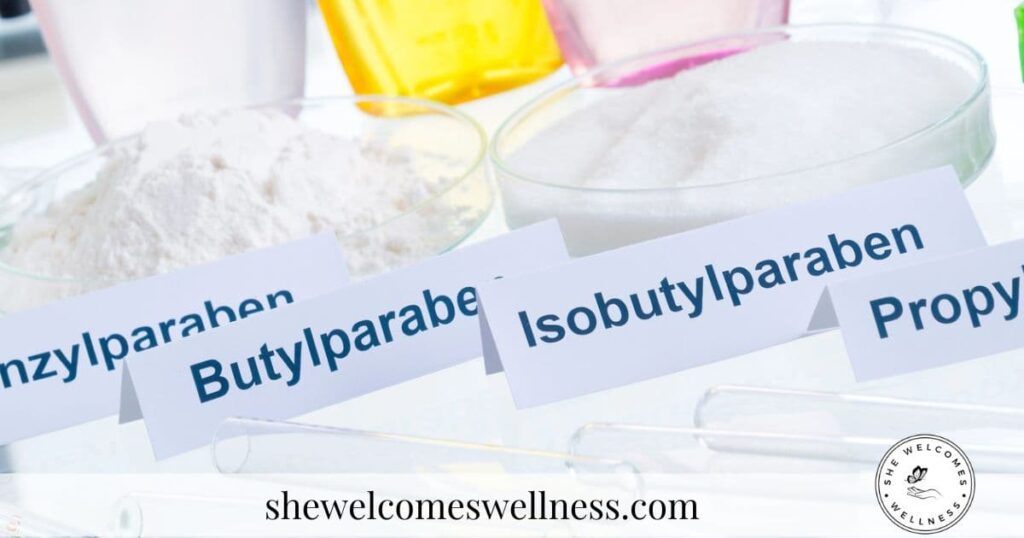 Close-up of glass dishes containing white powders labeled Benzylparaben, Butylparaben, and Isobutylparaben. Test tubes and colorful bottles in the background. Website "shewelcomeswellness.com" at the bottom.