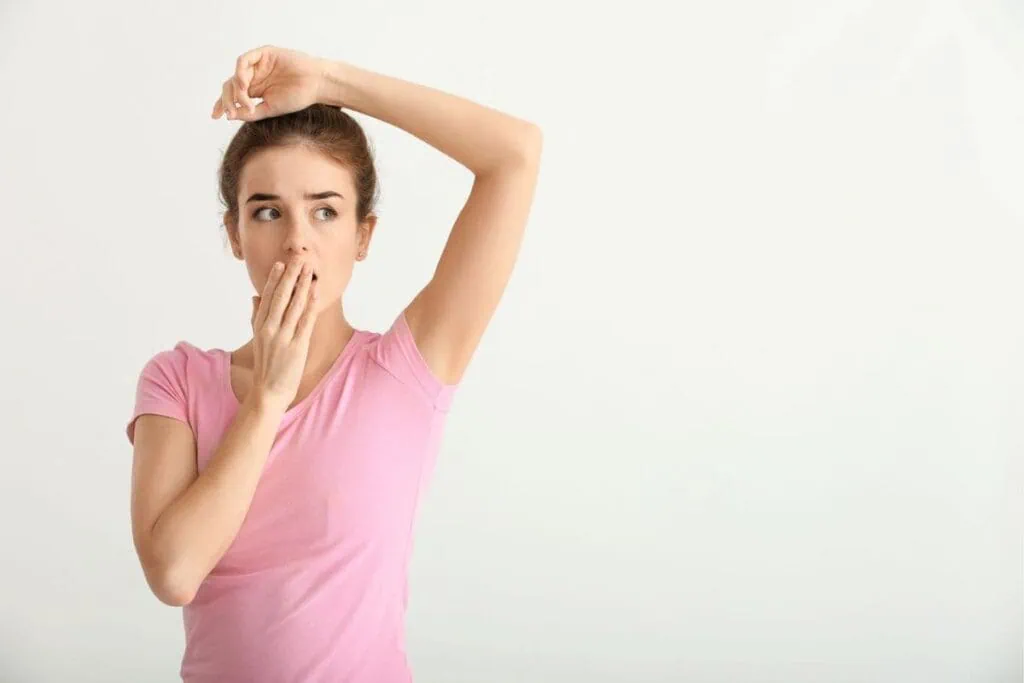 A woman in a pink shirt looks surprised, covering her mouth with one hand while raising the other arm, suggesting concerns about underarm odor. Concept for armpit detox and switching to natural deodorant.
