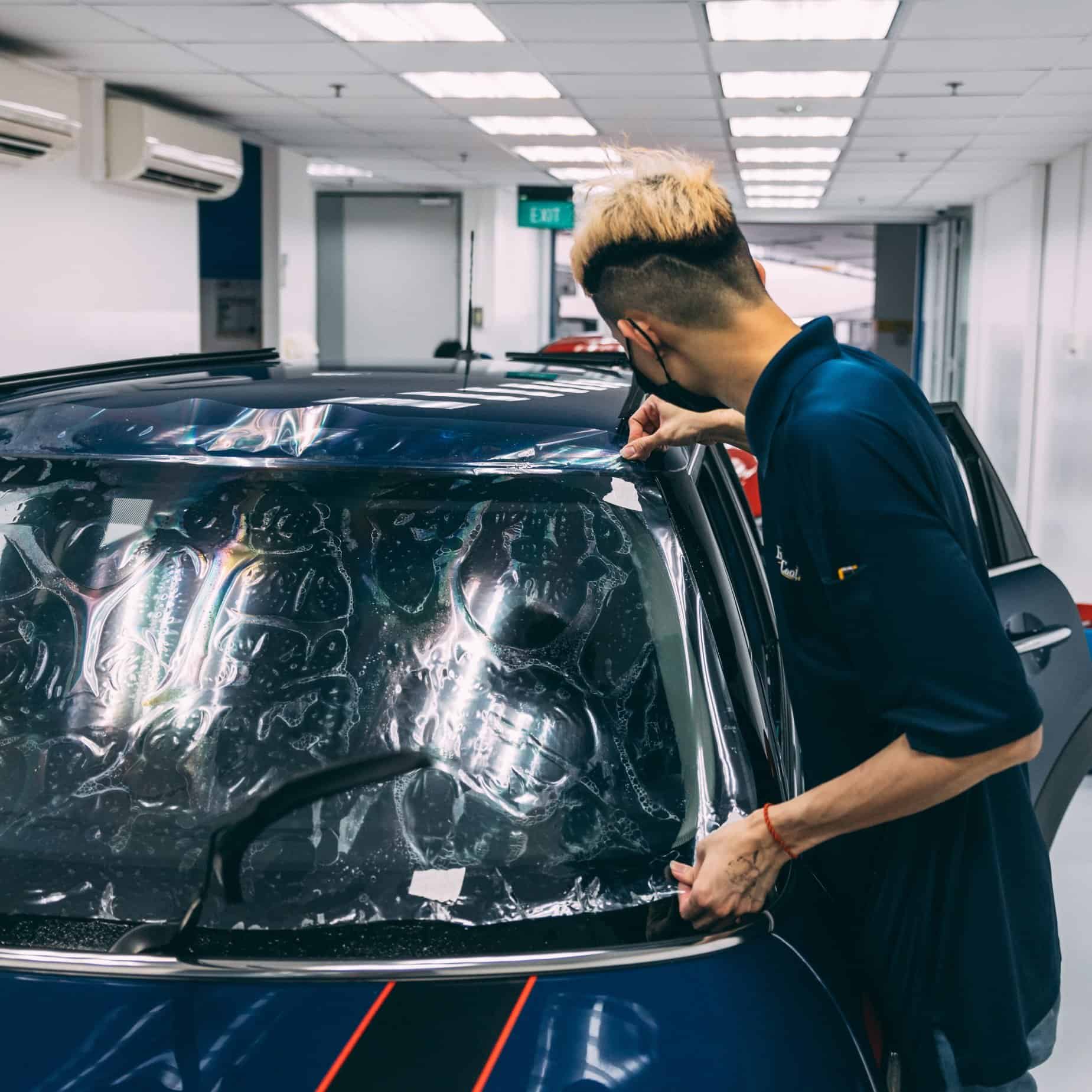 Technician applying stone chip protection film on car windshield and headlamp.