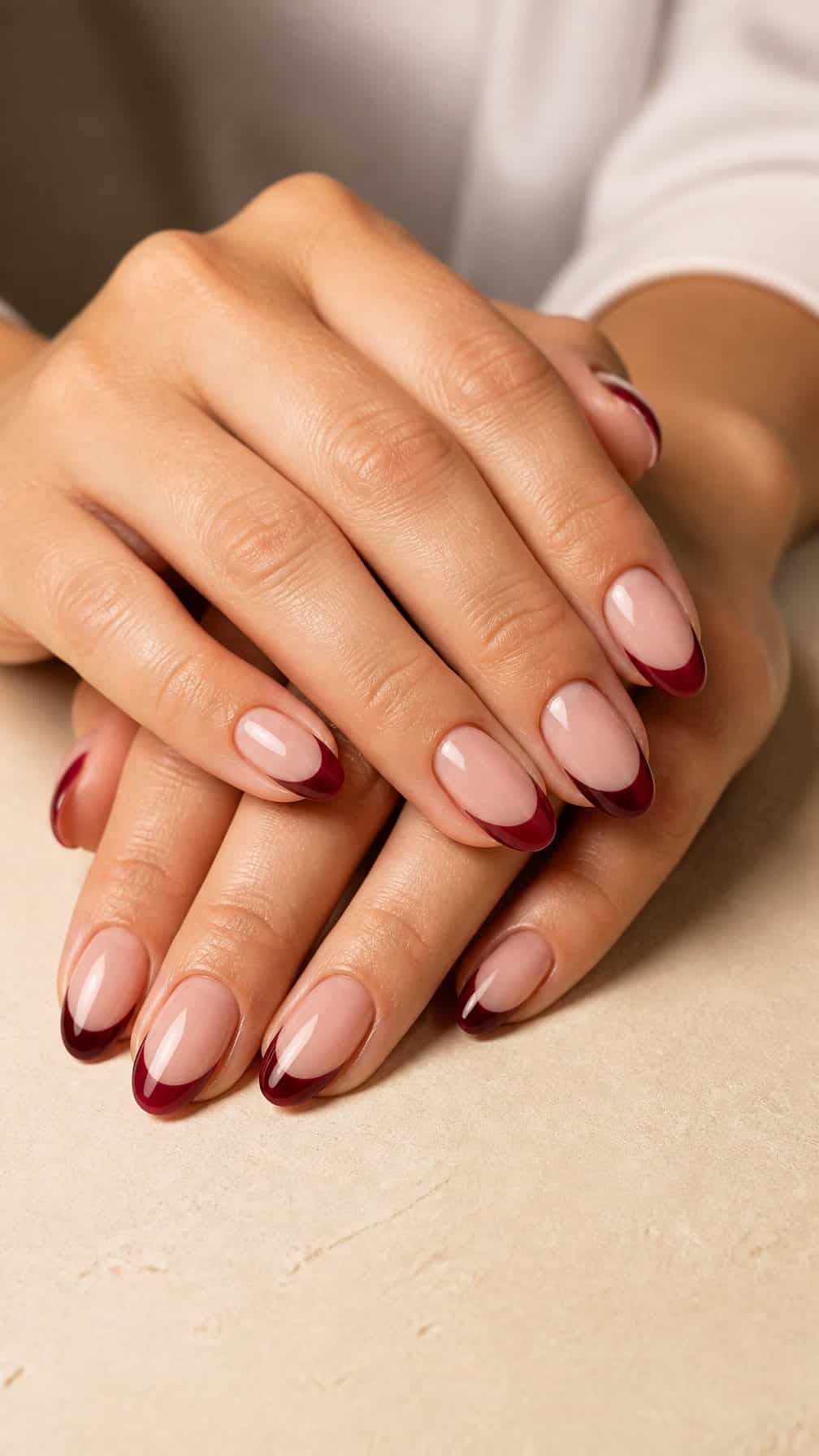 A close-up of manicured hands with glossy burgundy French-tip nails resting on a neutral surface.