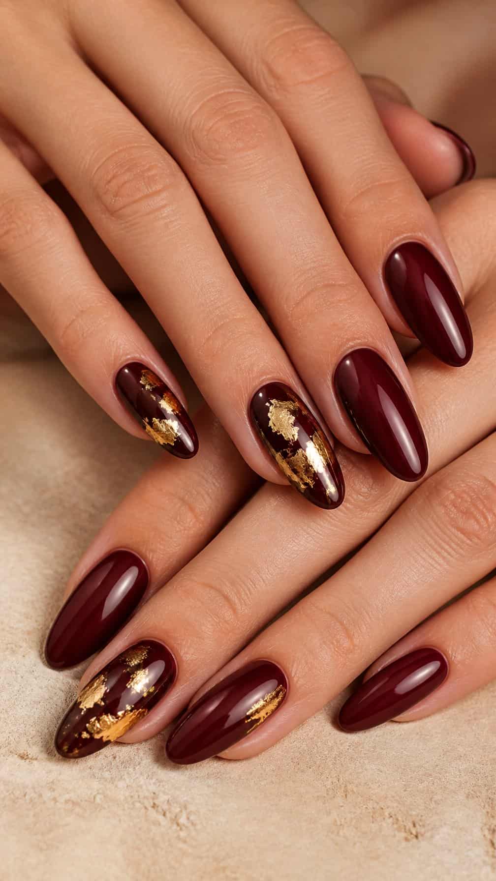 Close-up of a hand with glossy burgundy nails decorated with gold foil accents.