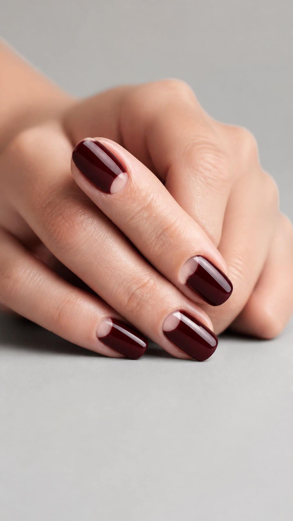 A hand with neatly manicured burgundy half-moon nails resting on a neutral surface.