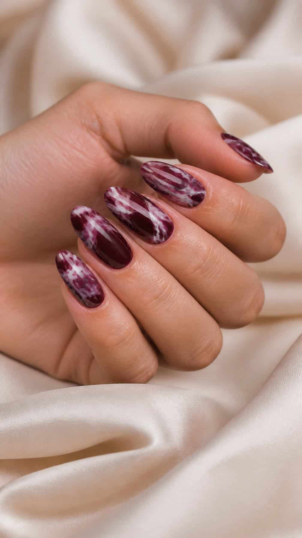 Close-up of a hand with glossy burgundy marble-patterned nails resting on a neutral surface.