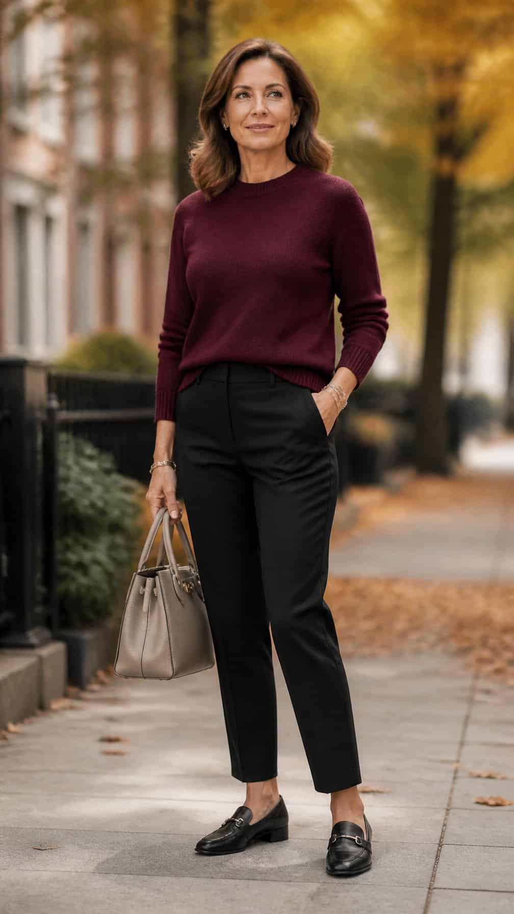 A mature woman wearing a burgundy sweater and black ankle pants stands outdoors among autumn foliage.