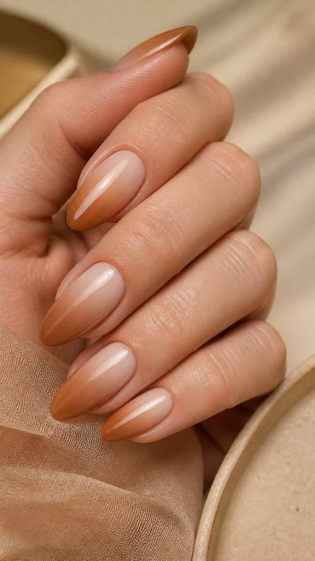 A hand displays glossy almond-shaped nails with a caramel-to-nude ombre manicure.