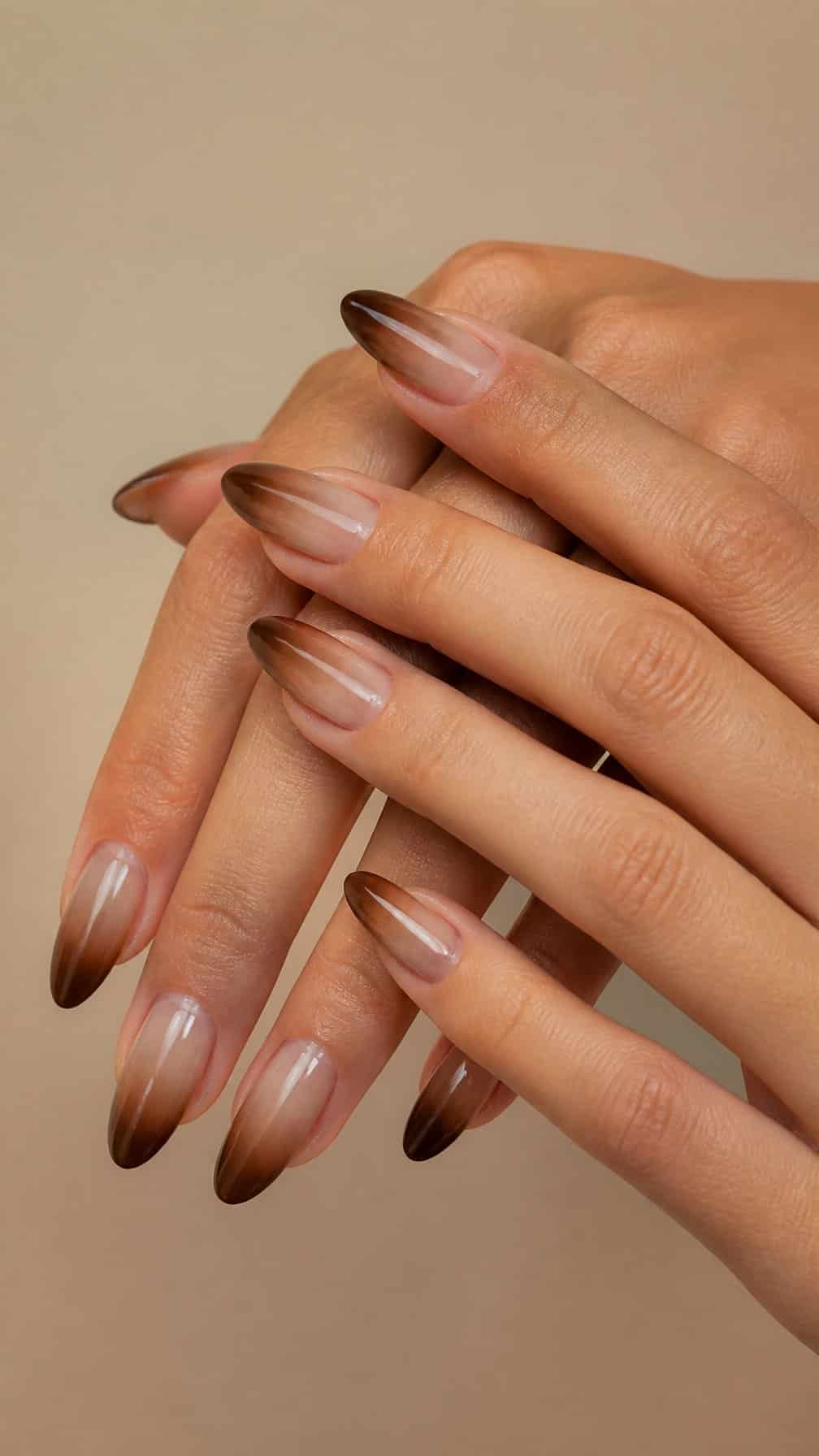 A close-up of hands with glossy cocoa brown to beige ombre almond-shaped nails.