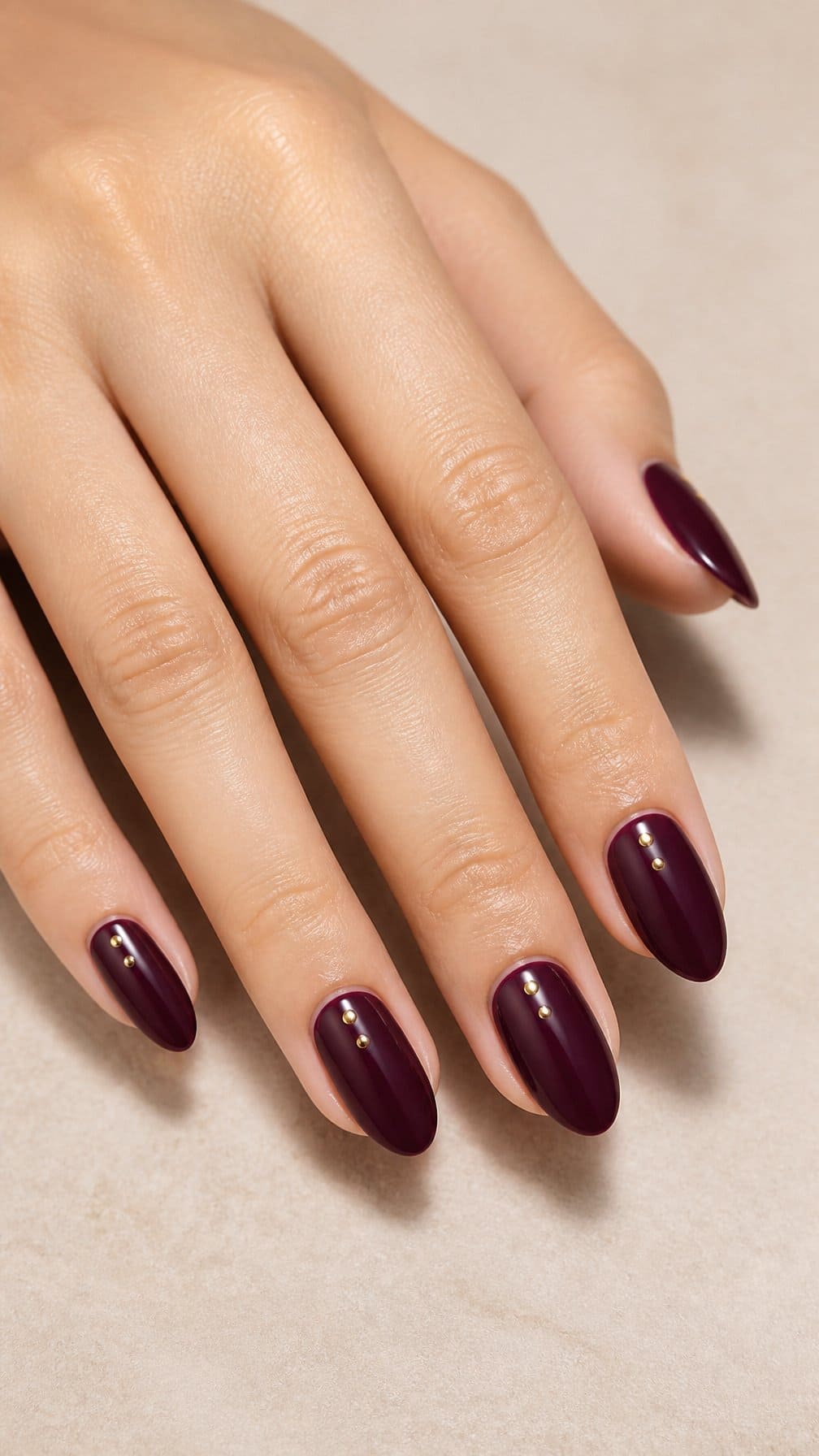 A hand with glossy deep plum nails decorated with small gold dots rests on a neutral surface.