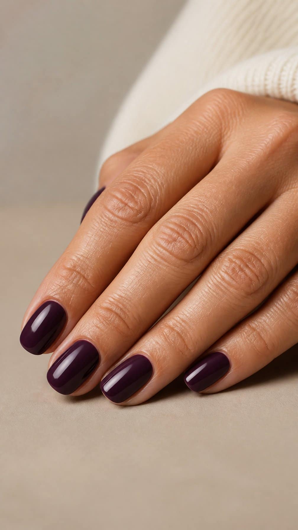 A mature woman’s hand with glossy deep plum oval-shaped nails resting on a neutral surface.