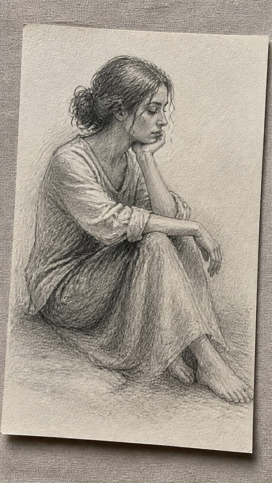 Hand-drawn contemplative seated figure in graphite drawing on textured paper, shown in a gentle oblique desk view that shows paper — study 1