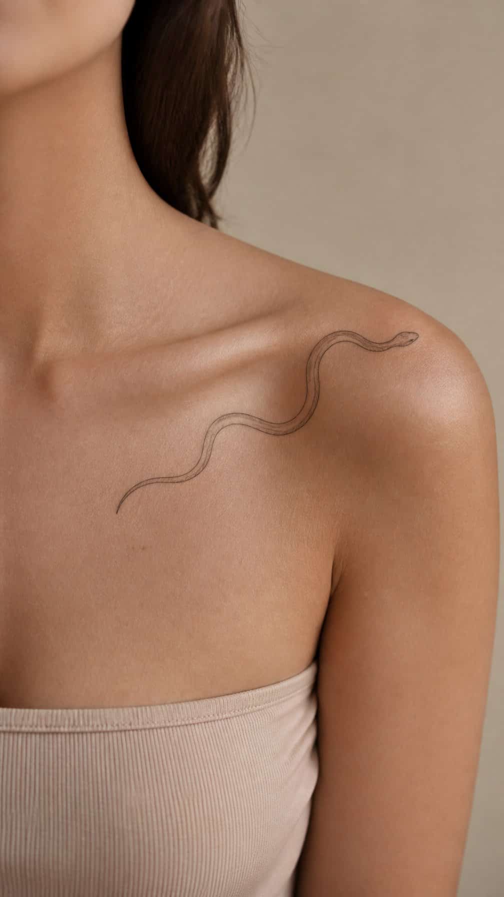 A fine-line snake tattoo curves from an adult model’s collarbone toward the shoulder.