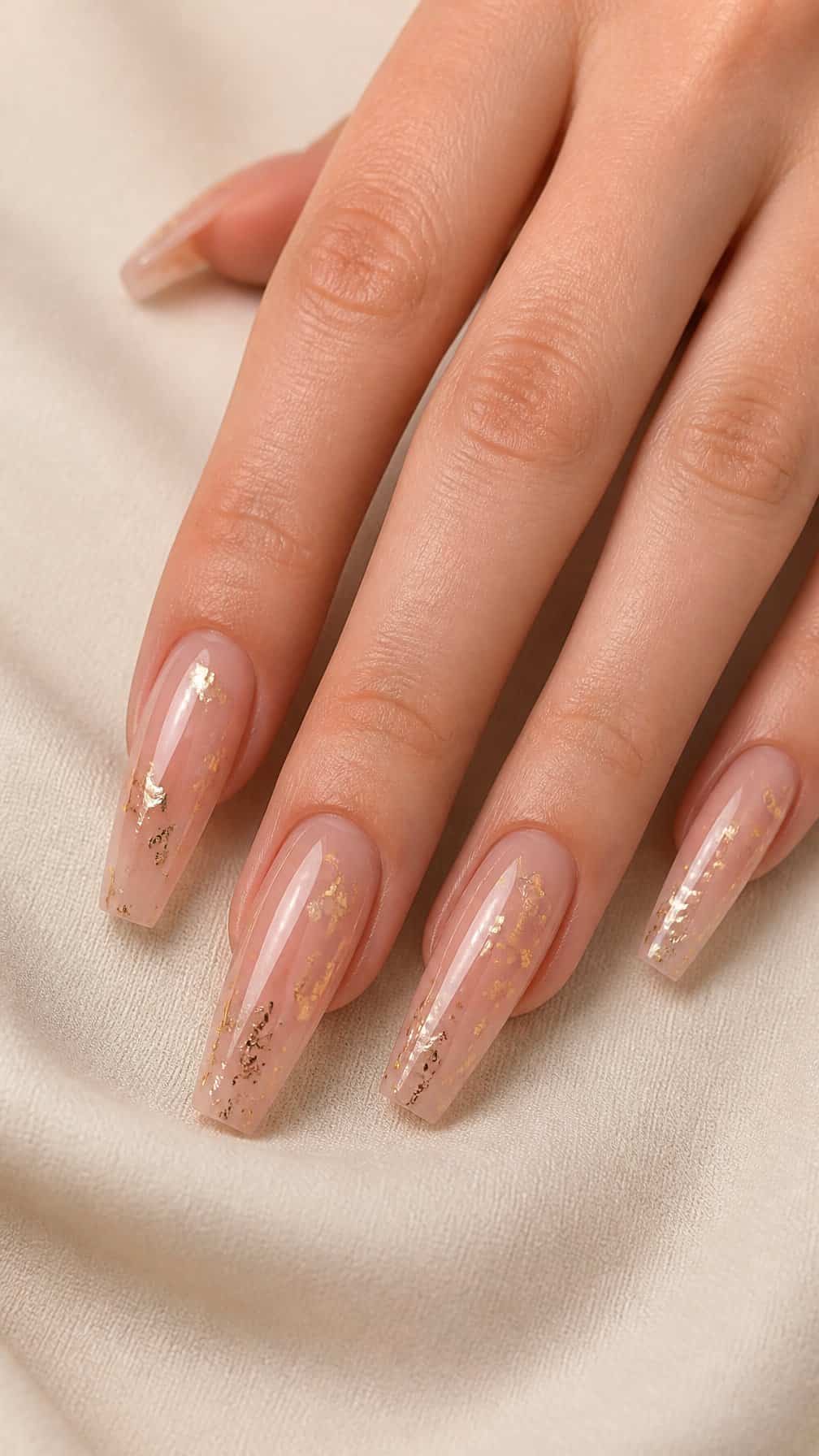 A hand displays long coffin-shaped nails with a nude base and gold flakes.