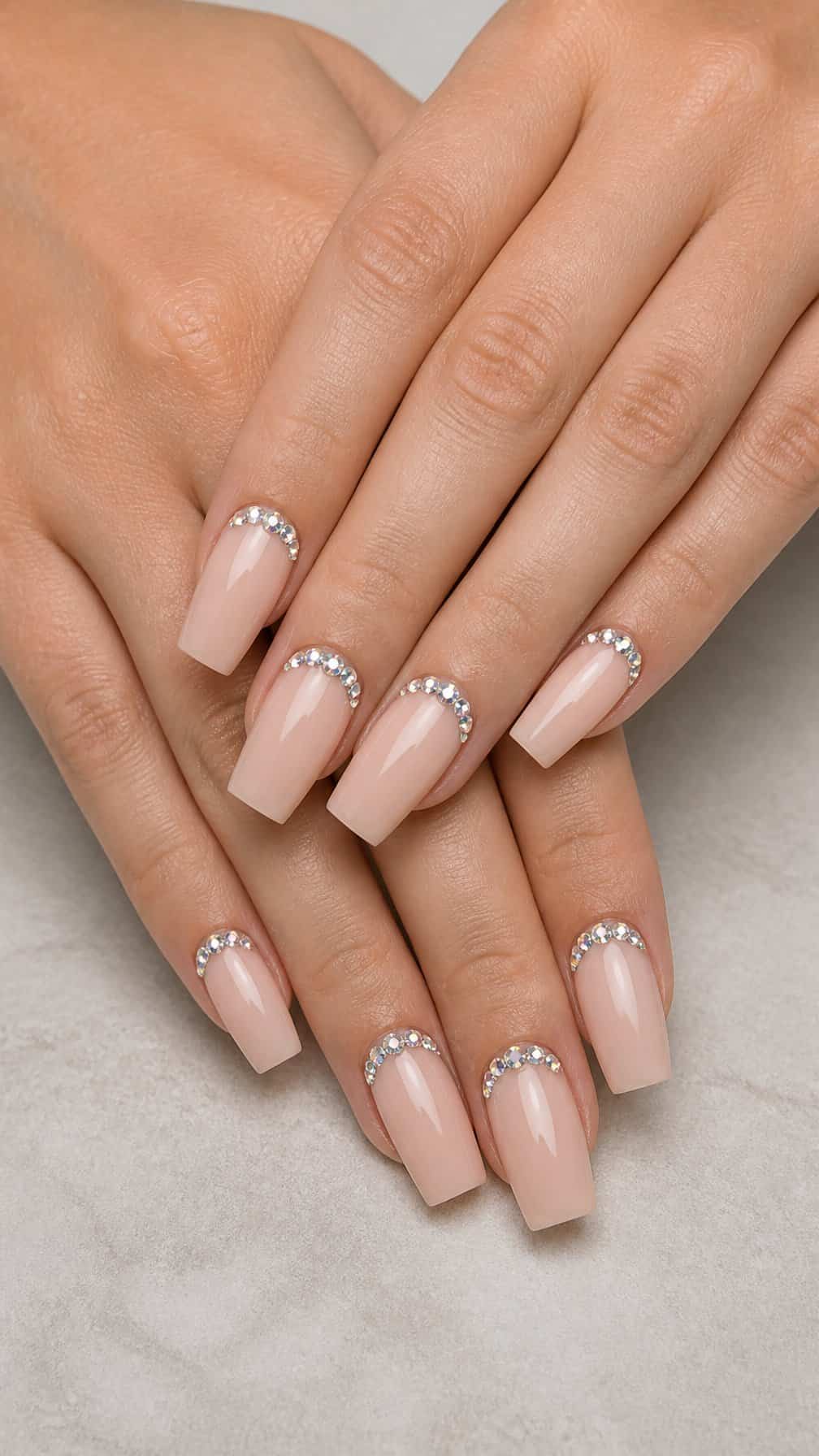 Close-up of long square nude nails decorated with rhinestones along the cuticles.