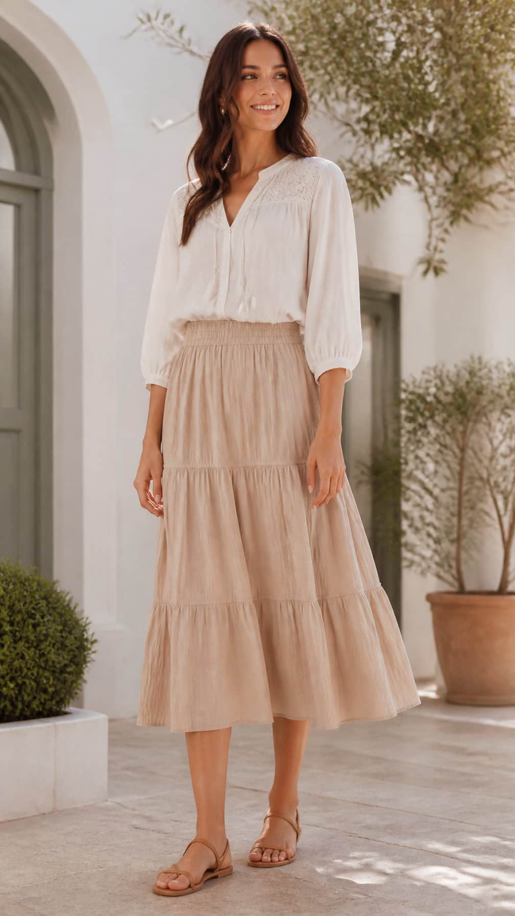 A woman stands outdoors wearing a tiered midi skirt, tucked-in blouse, and sandals.