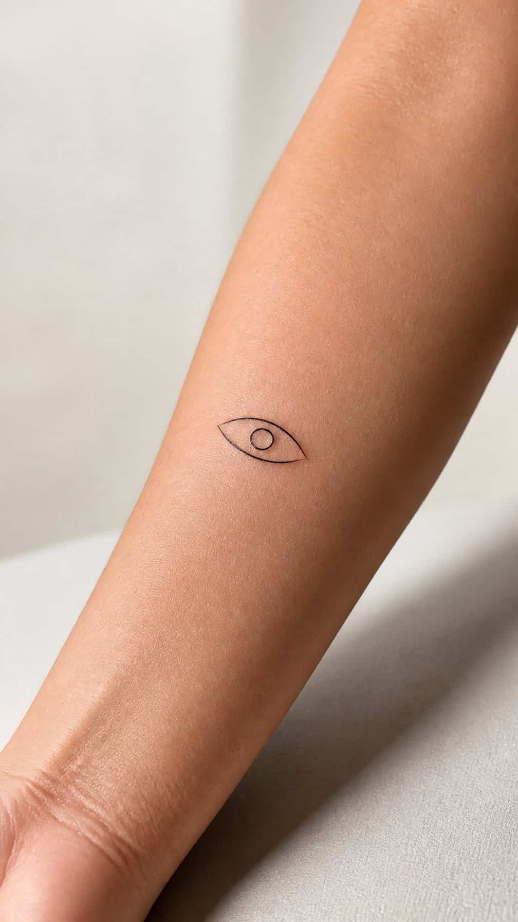 A minimalist eye tattoo on an adult person's inner forearm.