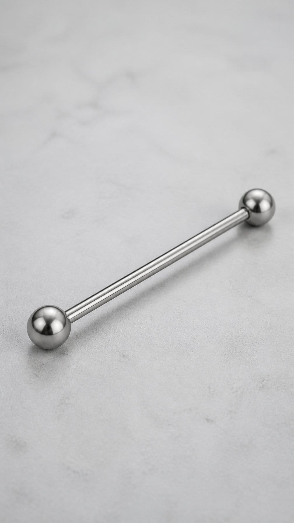 A minimalist titanium industrial barbell piercing rests diagonally on a neutral surface.