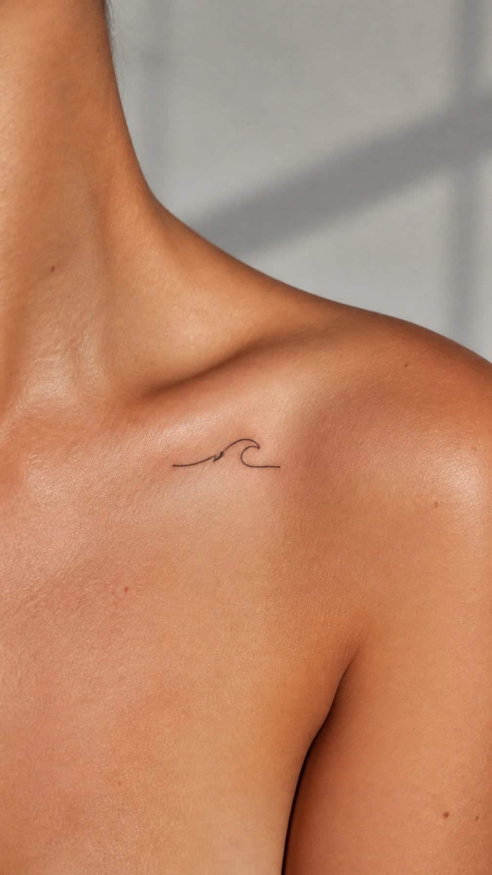 A small wave tattoo beneath an adult person’s collarbone near the shoulder.