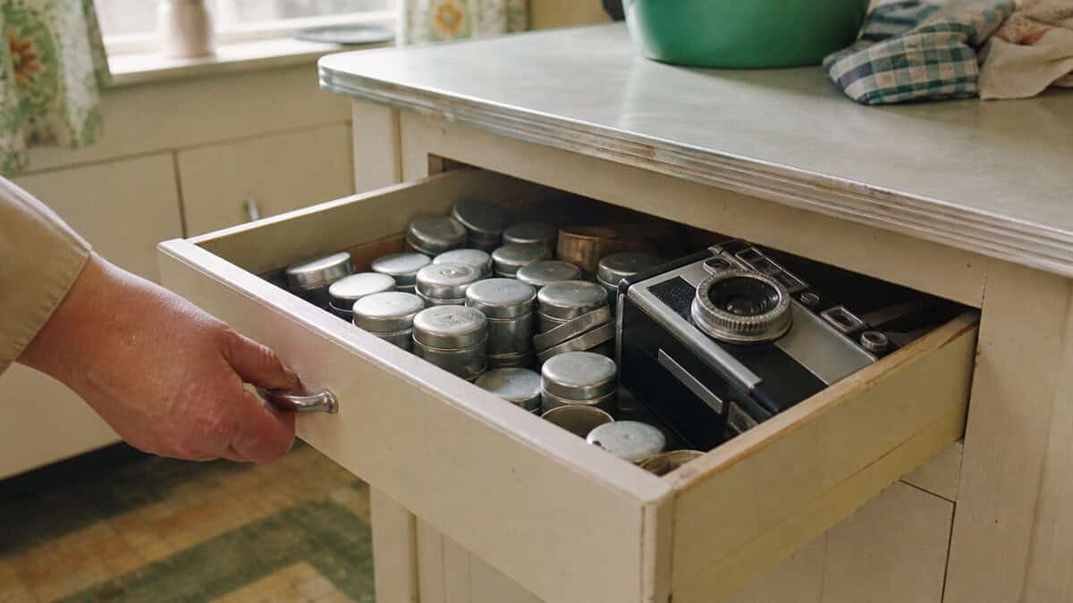 Hand opening a drawer of metal film canisters beside a compact 1960s camera