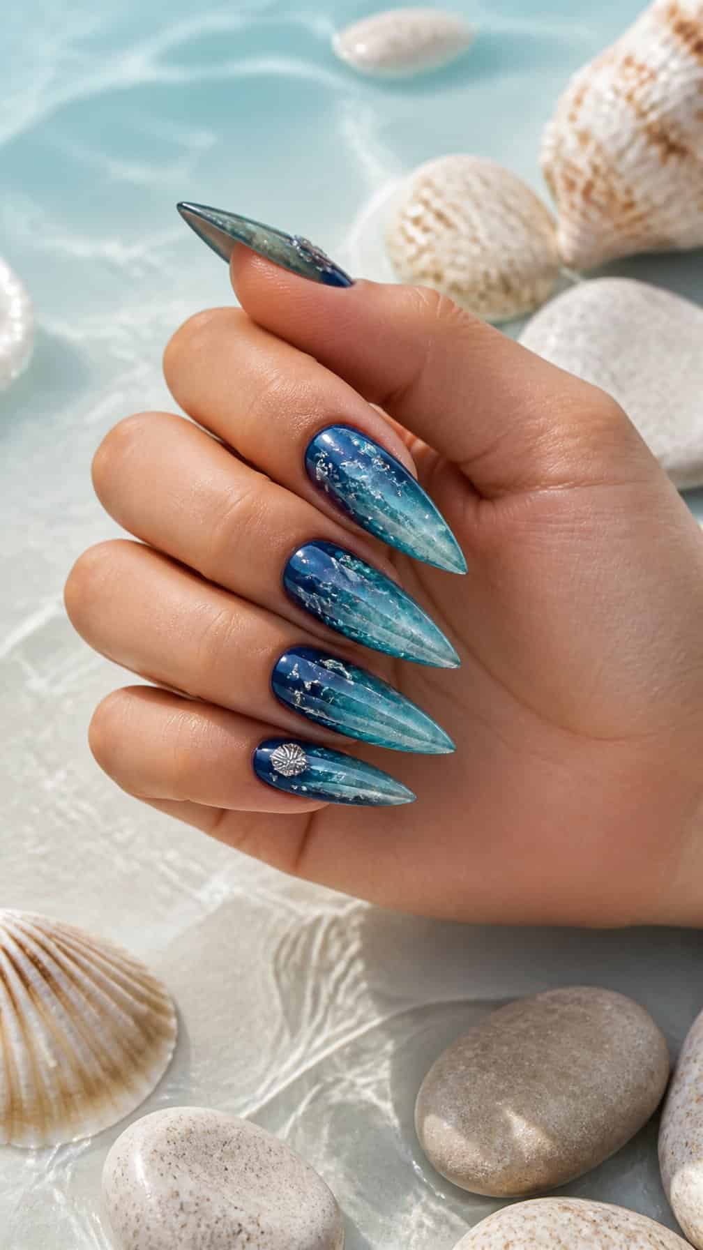 A hand displays long ocean-inspired blue stiletto nails beside seashells and smooth beach stones.